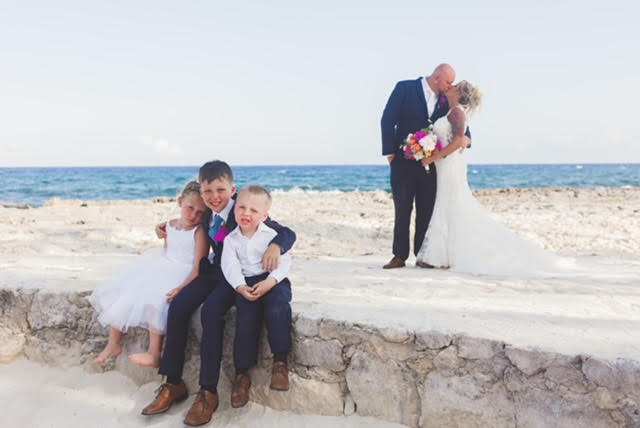 Bride and groom kissing on the beach with kids sitting in front of them.