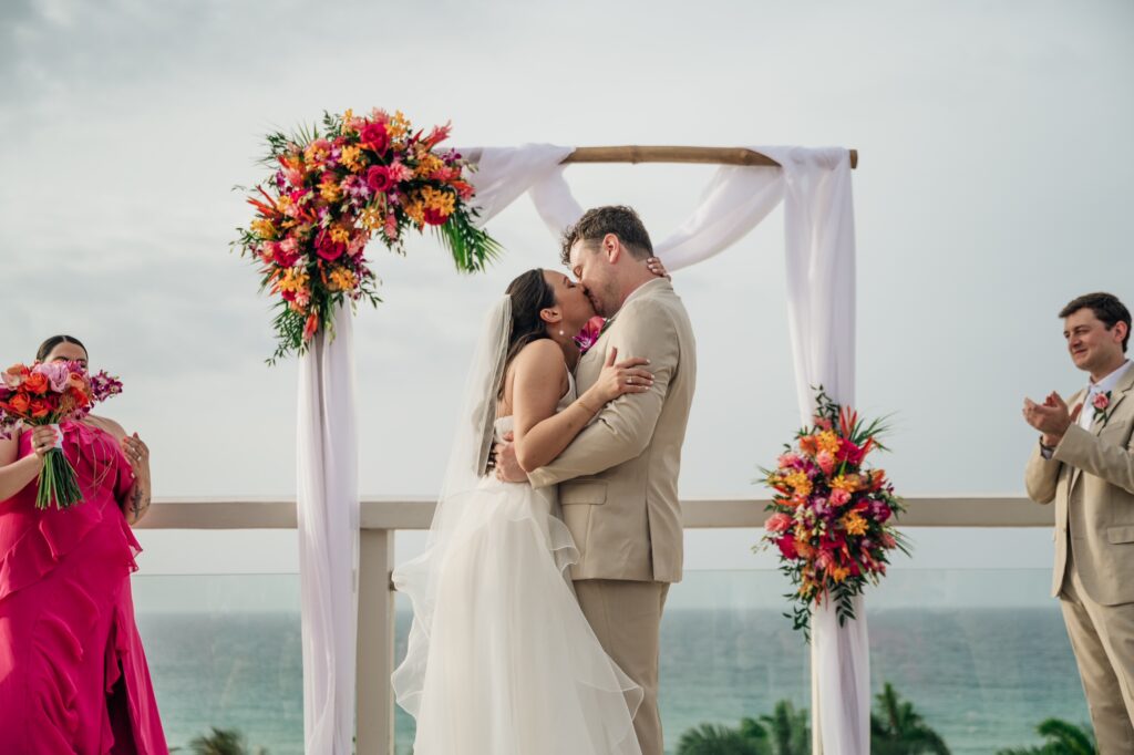 Wedding ceremony with bride and groom kissing under tropical flower arch